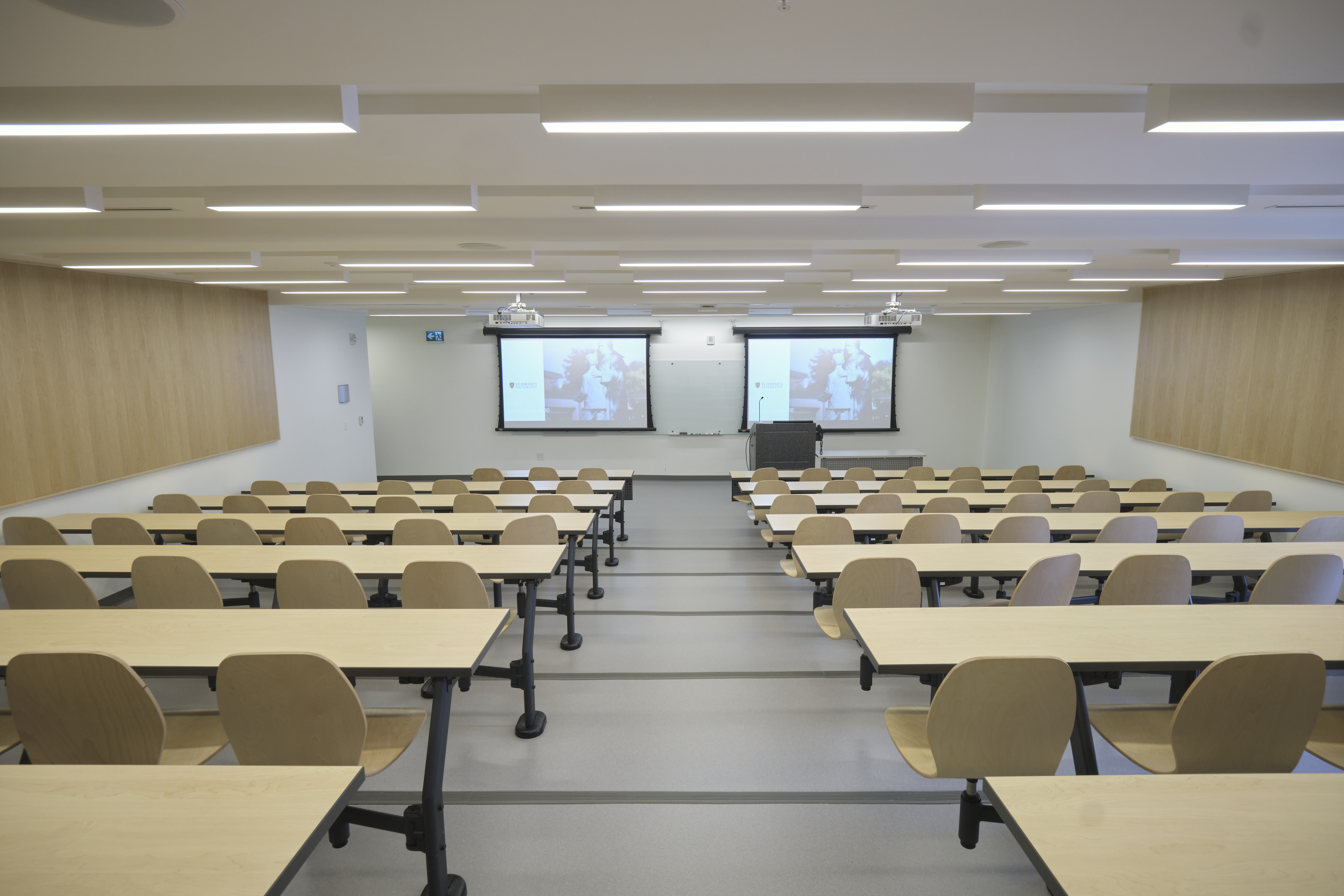 A classroom with fixed tiered seating and a lectern, whiteboards, and projector screens at the front.