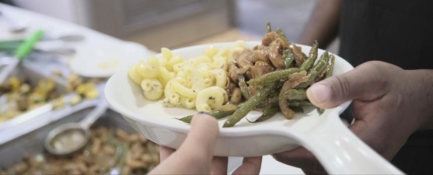 A food services staff hands a plate of pasta, meat and vegetables to a guest.