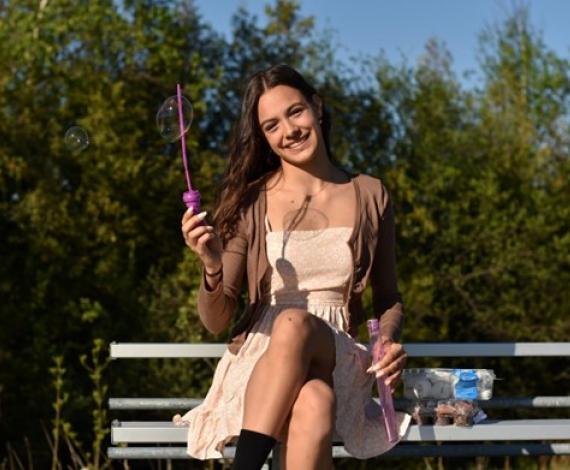 Ana, a femme presenting person, sits on a bench with a bubble wand, smiling.